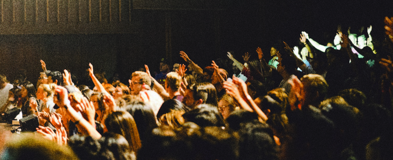 A side view of people in a lecture theatre. The lighting is dark and dramatic. The seats are tiered and everyone is facing an unseen speaker or screen on the left. People are engaged and many of those in the audience have their hands raised.